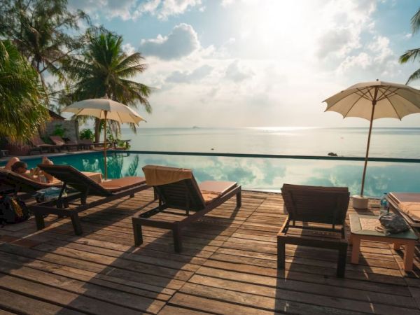 A sunlit deck with lounge chairs and umbrellas overlooks a serene infinity pool and ocean, surrounded by palm trees under a partly cloudy sky.