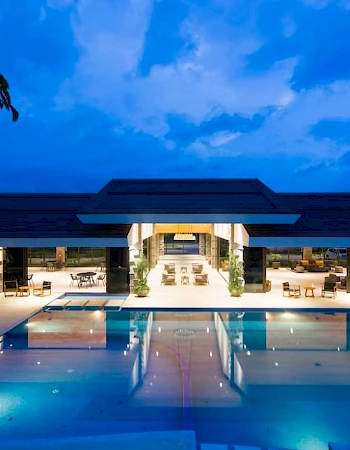 A luxurious modern building is reflected in a beautifully lit outdoor pool at twilight, surrounded by greenery and a spacious deck.