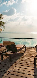 A tropical poolside scene with lounge chairs, umbrellas, and palm trees under a sunny sky near the ocean.