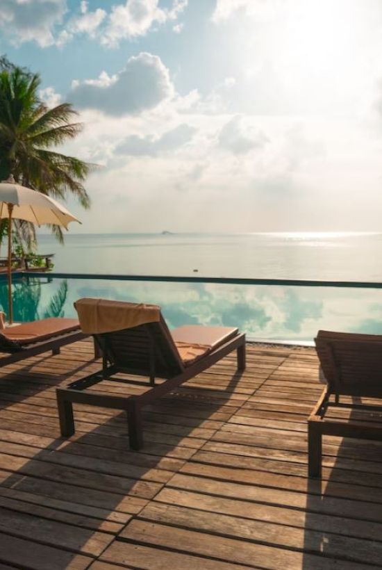 A serene seaside pool deck with wooden loungers, umbrellas, and a view of the calm sea under a partly cloudy sky.