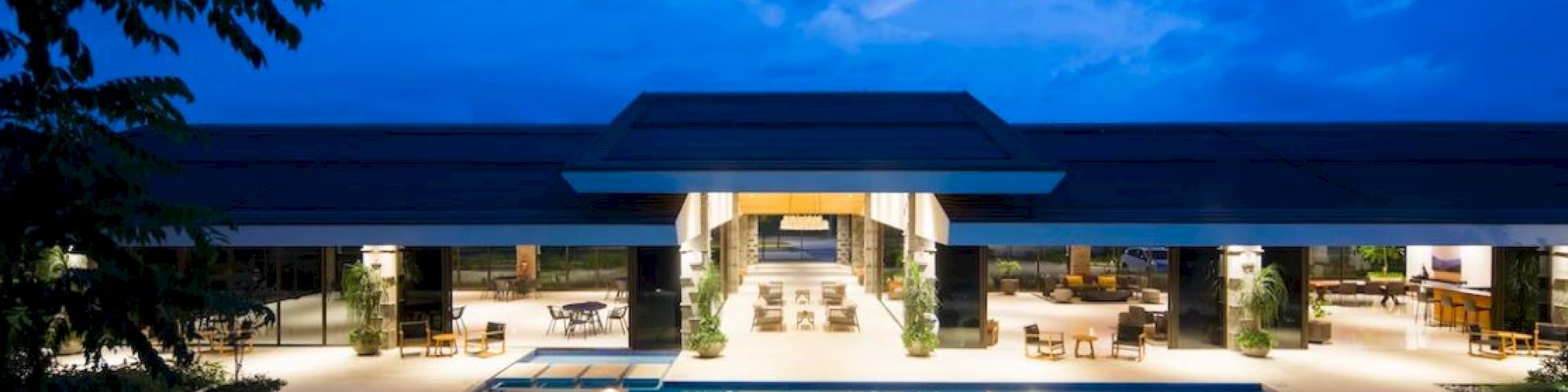The image shows a modern building with an illuminated pool, surrounded by trees, set against a twilight sky.