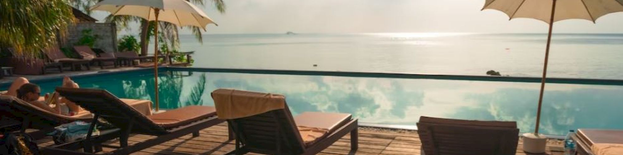 A tranquil poolside scene with lounge chairs, parasols, palm trees, and a view of the calm sea beneath a cloudy sky.