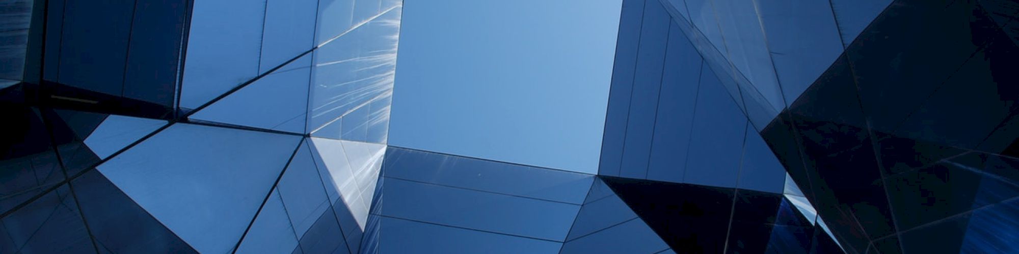 Geometric architectural shapes form an abstract pattern with reflective surfaces, creating a view of the sky through angular structures.