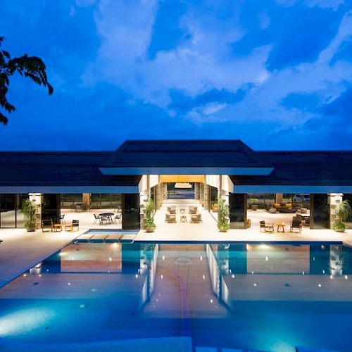 The image shows a luxurious, modern building with a large, illuminated swimming pool in the foreground, under a twilight sky.