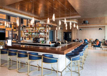 It’s a modern bar with a central counter, stylish seating, and pendant lighting, creating an inviting atmosphere.