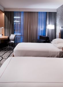 The image shows a hotel room with two beds, a flat-screen TV, a work desk, and modern lighting, featuring a stylish decor.
