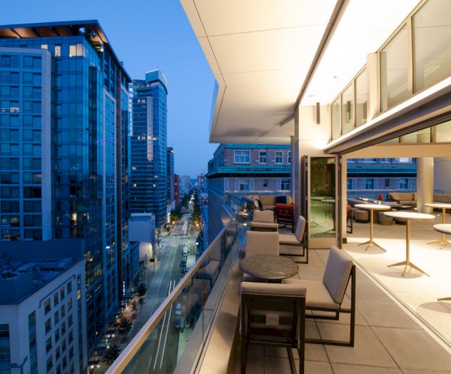A rooftop terrace with seating overlooks a cityscape at dusk, featuring high-rise buildings and a stylish, illuminated outdoor area.