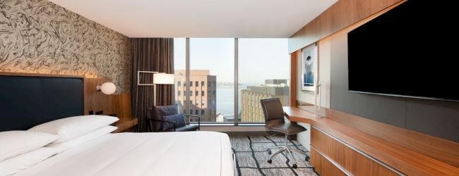 Modern hotel room with a large bed, wall-mounted TV, desk, chair, and a window offering a city view.