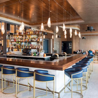A modern bar with a central counter, blue stools, and hanging lights in a stylish setting.
