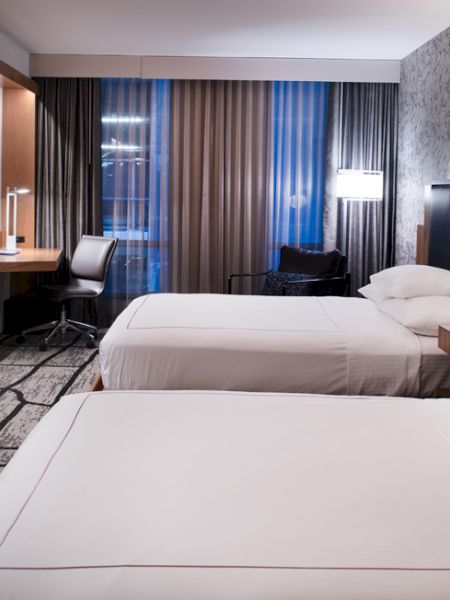 The image shows a modern hotel room with two beds, a flat-screen TV, a desk with a chair, and a window with curtains, in neutral tones.