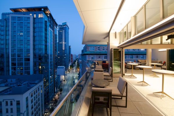 A modern rooftop terrace with seating, overlooking a cityscape with tall buildings and a street below at dusk.