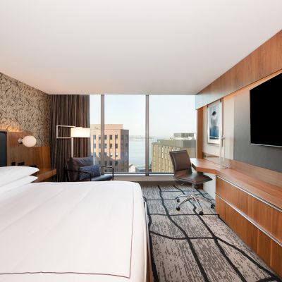 A modern hotel room with a large bed, wall-mounted TV, desk, and a window offering a view of buildings outside.