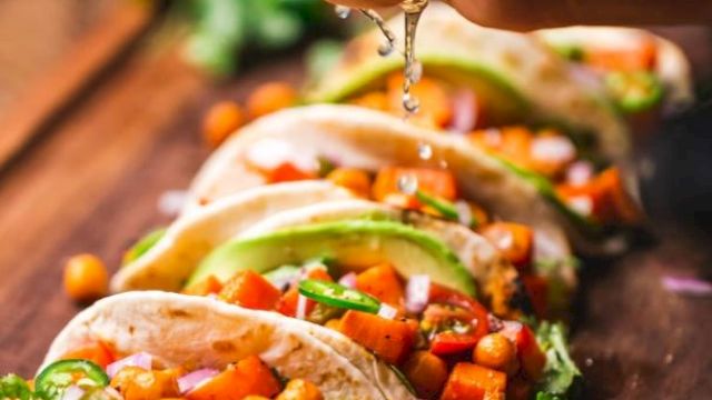 The image shows vibrant tacos with chickpeas, avocado, onions, and a hand squeezing lime juice over them on a wooden board.