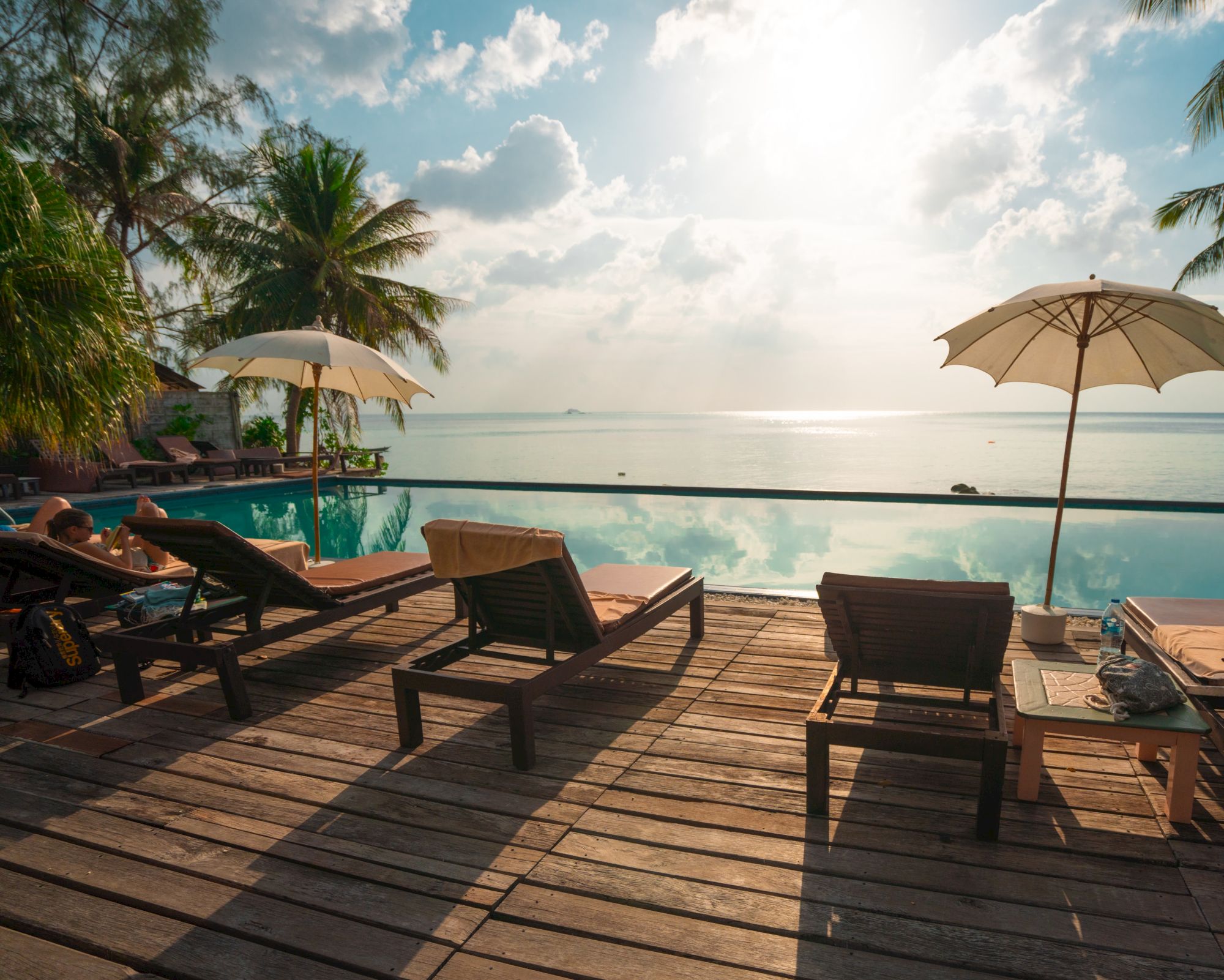 The image shows a serene poolside setting with lounge chairs, umbrellas, and a view of the sea under a partly cloudy sky.