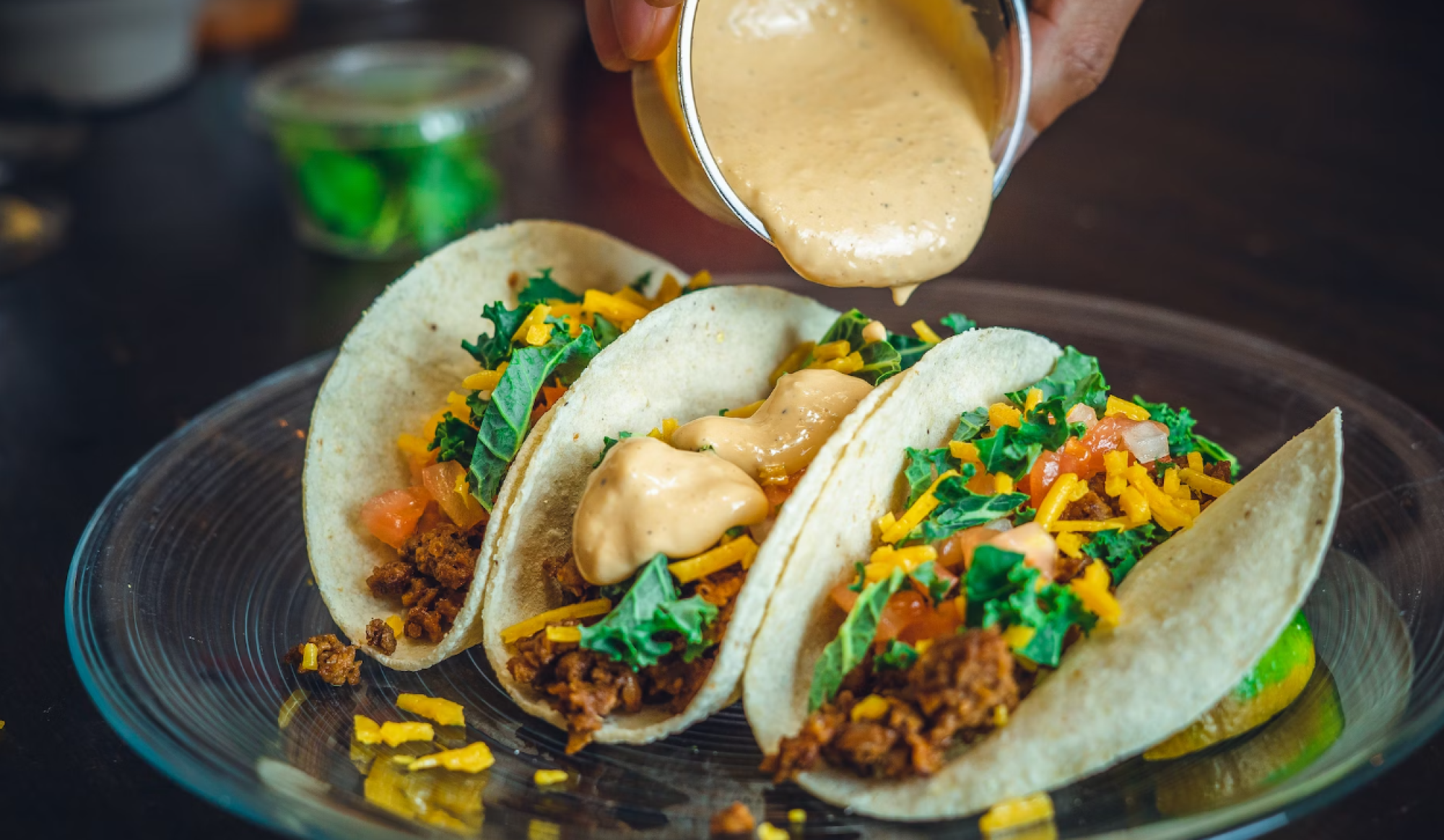 Three tacos filled with meat, cheese, tomatoes, and kale being topped with sauce on a glass plate.