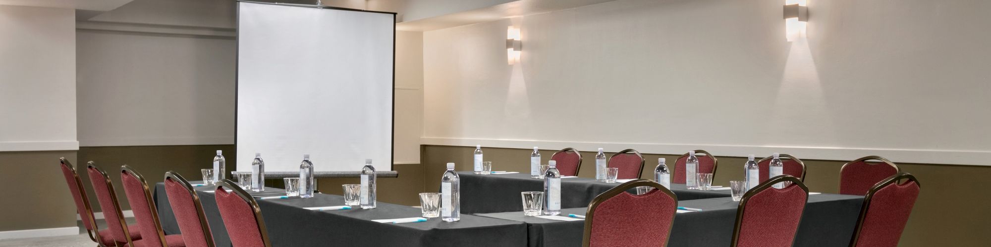 A conference room with U-shaped table setup, red chairs, water bottles, and a projection screen in a neutral-toned space.