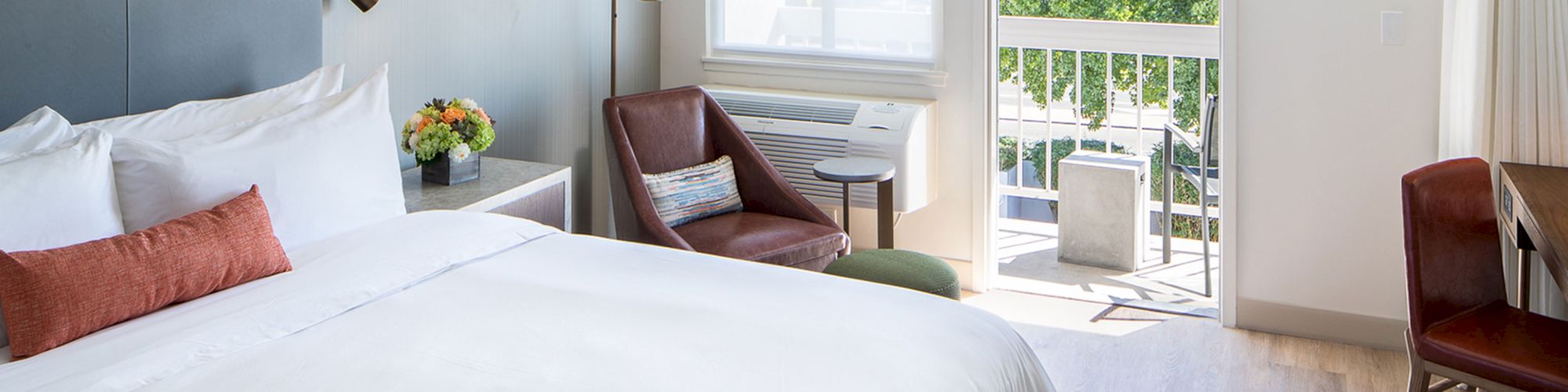 The image shows a cozy bedroom with a white bed, brown chair, desk, window, flowers, and a balcony with a view outside.