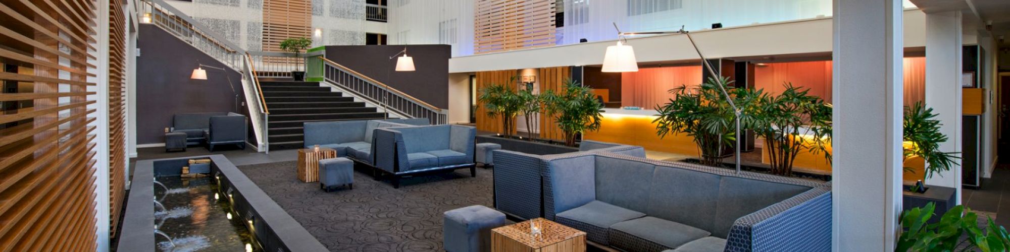 A modern lounge area with gray seating, wooden tables, plants, and a water feature, under a high ceiling with a staircase.