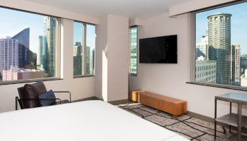 A modern hotel room with large windows showcasing a cityscape view, a wall-mounted TV, a bed, a chair, and a small table near the window.