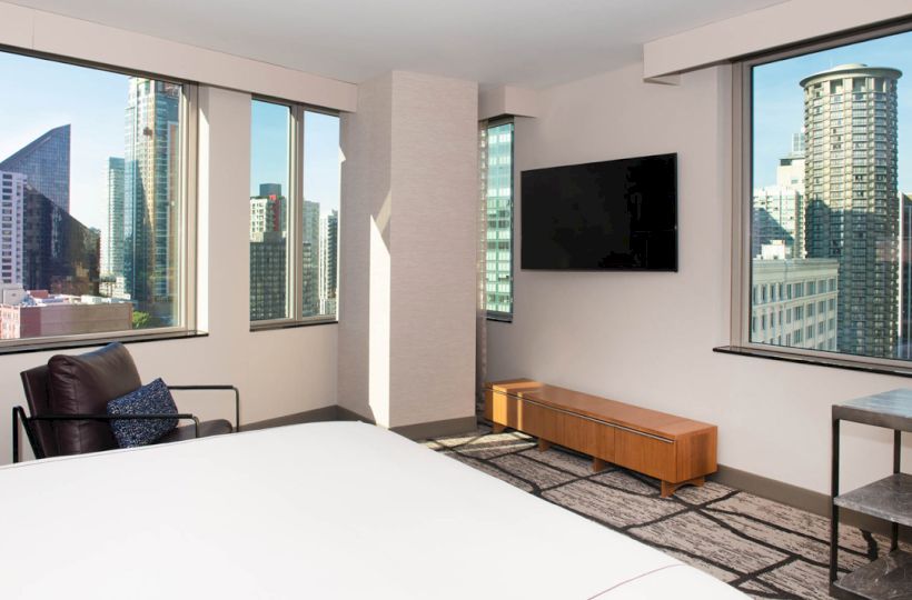 A modern hotel room with large windows showcasing a cityscape view, a wall-mounted TV, a bed, a chair, and a small table near the window.