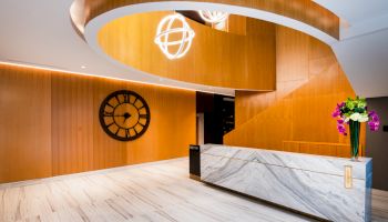 A modern lobby with a marble reception desk, wooden paneling, a clock on the wall, and a decorative light fixture above.