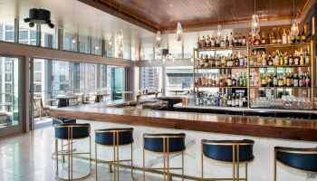 The image shows a modern bar with high stools, a stocked bar shelf, and city views through large windows, creating a stylish ambiance.