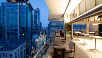 A modern rooftop terrace with outdoor seating overlooks a city street lined with tall buildings, captured at dusk.