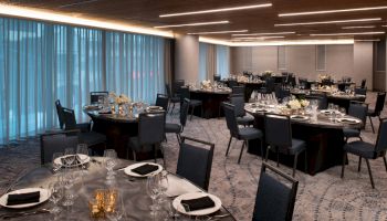 A banquet hall is set up with round tables covered with plates, glasses, and dark napkins, under ambient lighting.