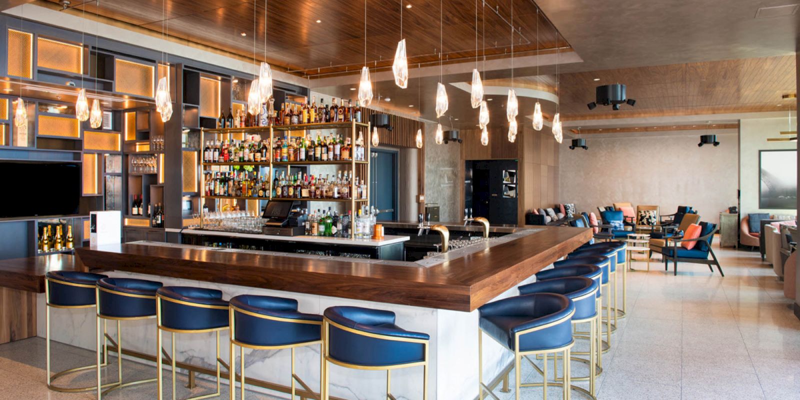 The image shows a stylish bar with blue stools, a wooden counter, hanging lights, and a well-stocked back bar, creating a modern ambiance.