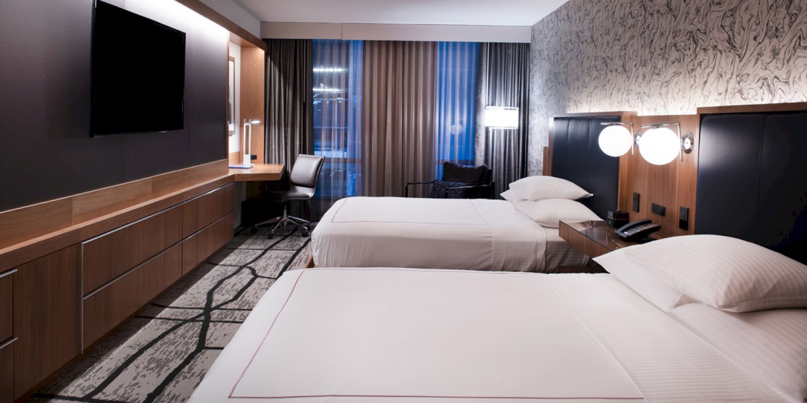 A hotel room with two beds, a TV, a desk, lamps, and curtains. The decor is modern and minimalistic, with soft lighting and neutral tones.