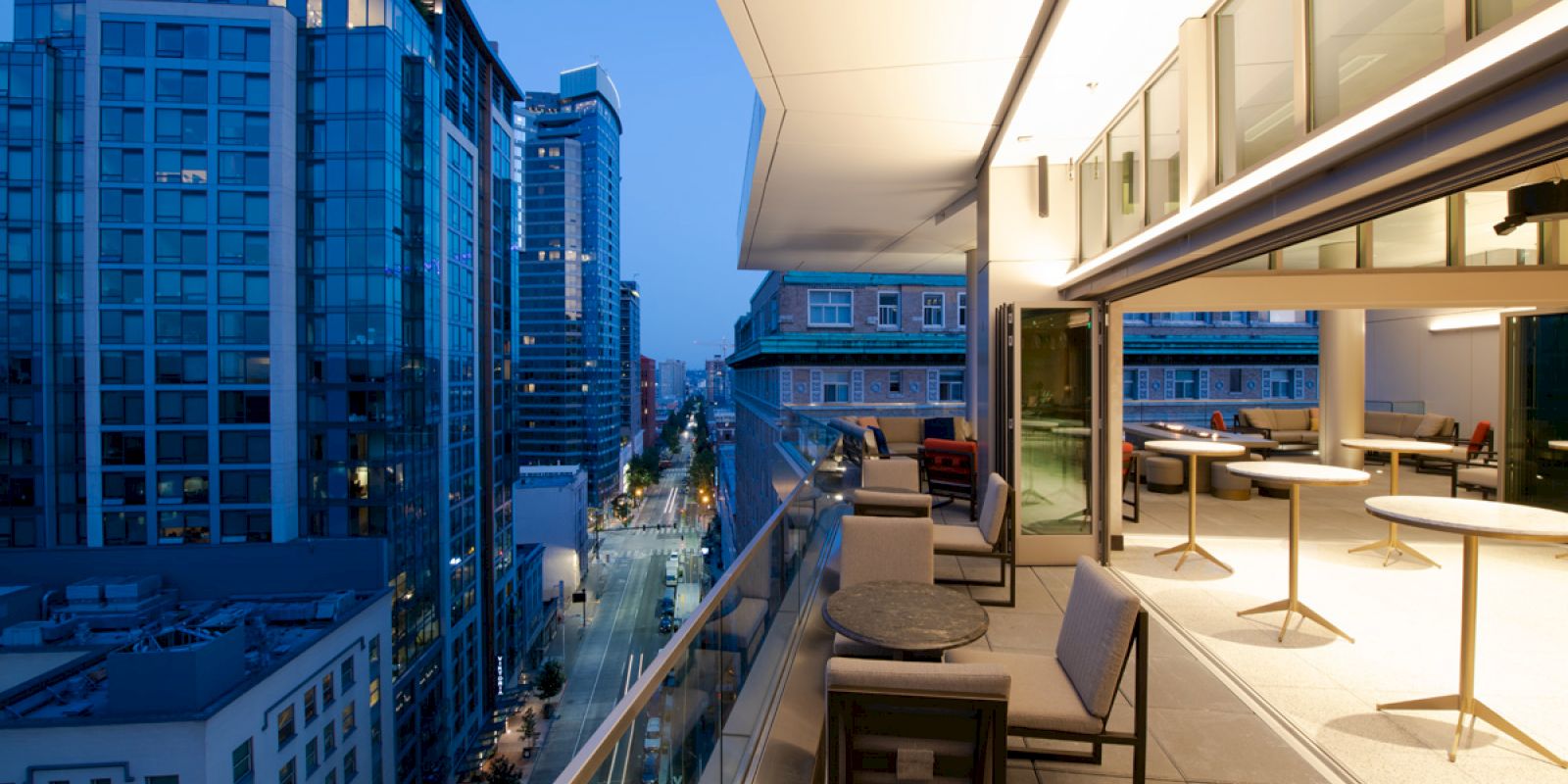A modern balcony overlooking a cityscape at dusk, featuring patio seating and glass buildings in the background.