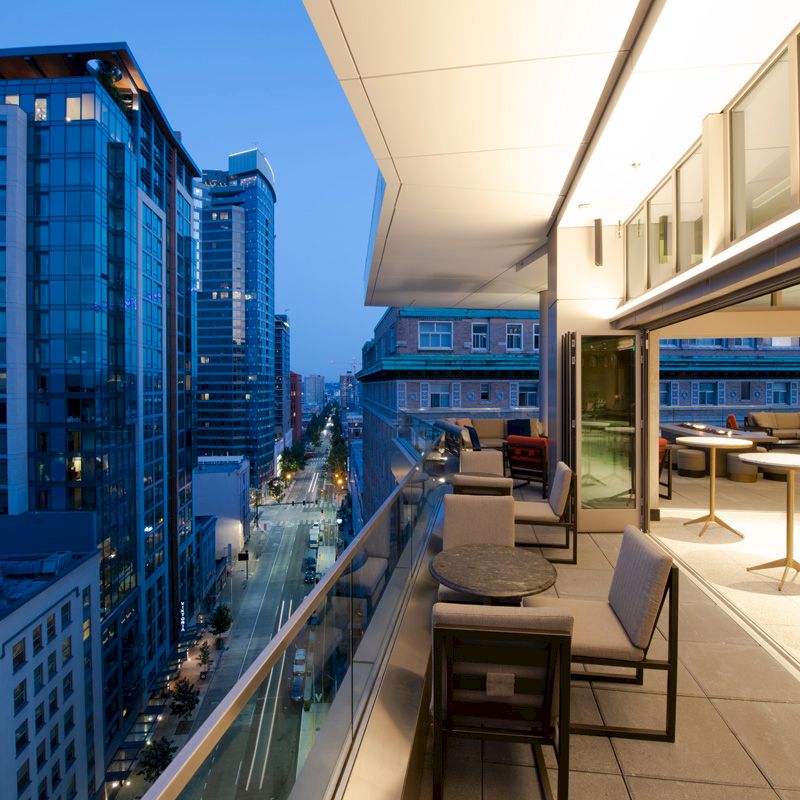 A rooftop terrace with modern seating overlooks a cityscape at dusk, featuring tall buildings and a lively, illuminated street below.