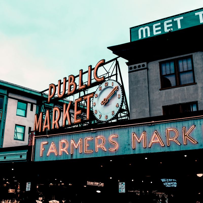 The image shows a neon sign for a public and farmers market, with a clock and nearby buildings under a cloudy sky.