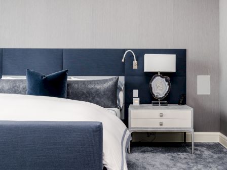 A modern bedroom with a blue headboard, white bedding, a nightstand with a lamp, and a decorative mirror on a grey carpet.