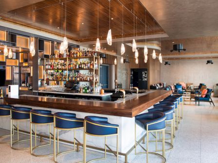 The image shows a stylish, modern bar with blue stools, a well-stocked liquor shelf, and decorative lighting in a spacious setting.