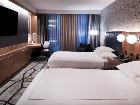 A modern hotel room with two beds, a TV, desk, chair, and lamps, featuring neutral colors and patterned carpeting.