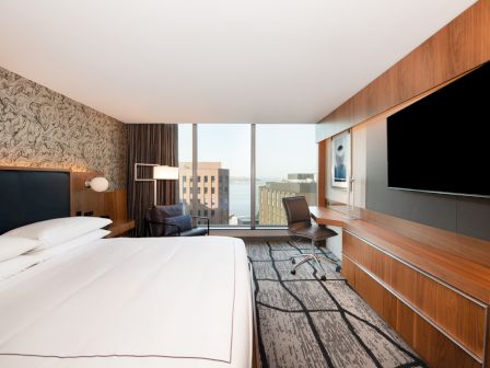 The image shows a modern hotel room with a bed, large TV, desk, chair, and floor-to-ceiling window with a city view.