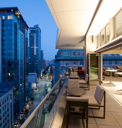 A modern balcony with tables and chairs overlooks a cityscape at dusk, featuring tall buildings and a well-lit street below.
