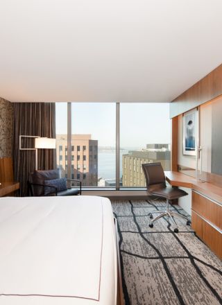 A modern hotel room with a large bed, a desk, a chair, and a flat-screen TV, featuring a window view of city buildings.
