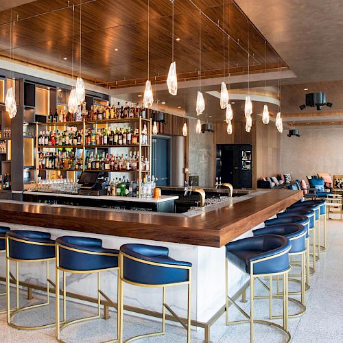 The image shows a modern bar with blue chairs, pendant lights, and assorted bottles on shelves, creating a cozy and stylish ambiance.