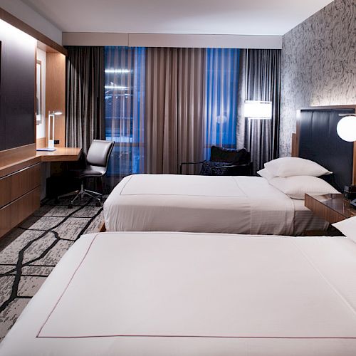 The image shows a modern hotel room with two beds, a wall-mounted TV, a desk, and stylish lighting.