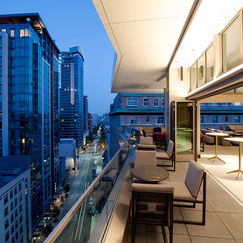 A modern outdoor terrace with seating overlooks a city street lined with tall buildings at dusk.