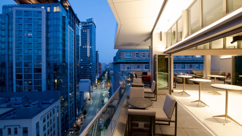 A rooftop patio with modern seating overlooks a cityscape of illuminated skyscrapers and streets during twilight.