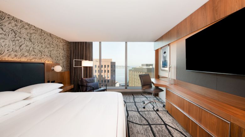 A modern hotel room with a large bed, a wall-mounted TV, and a window view of buildings and cityscape.