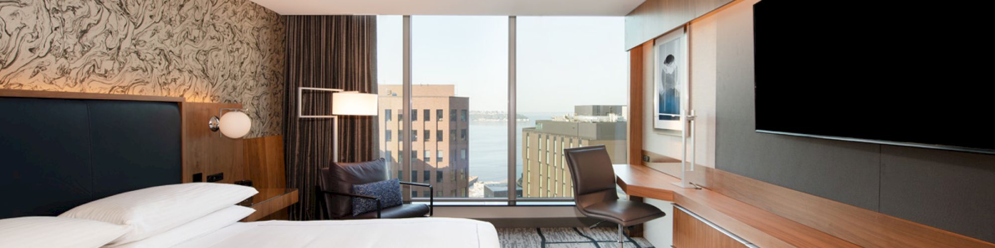 A modern hotel room with a large bed, desk, chair, flat-screen TV, and floor-to-ceiling windows with a city view.